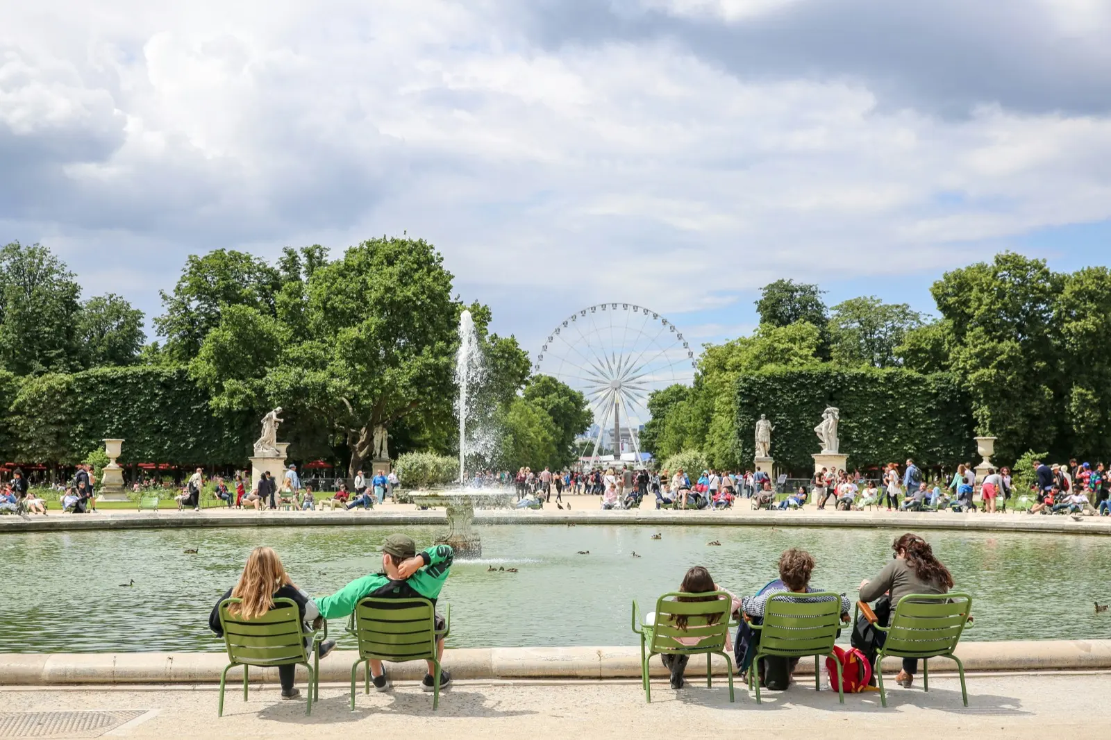 Des promeneurs assis sur des chaises vertes au bord d’un bassin avec jet d’eau au jardin des Tuileries à Paris, entouré de statues et d’arbres, avec une grande roue en arrière-plan.
