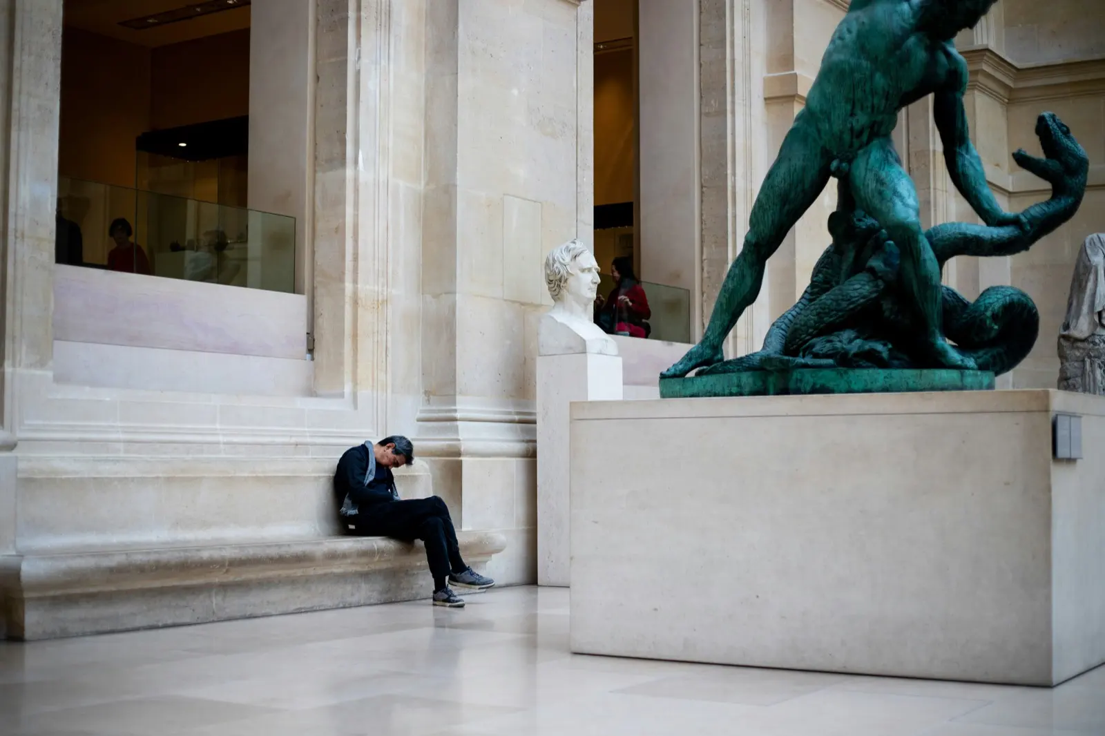 Dans une grande salle de musée en pierre claire, un homme assis sur un banc, tête baissée, à côté d’un buste en marbre et d’une statue de bronze verte représentant un homme luttant contre un serpent.