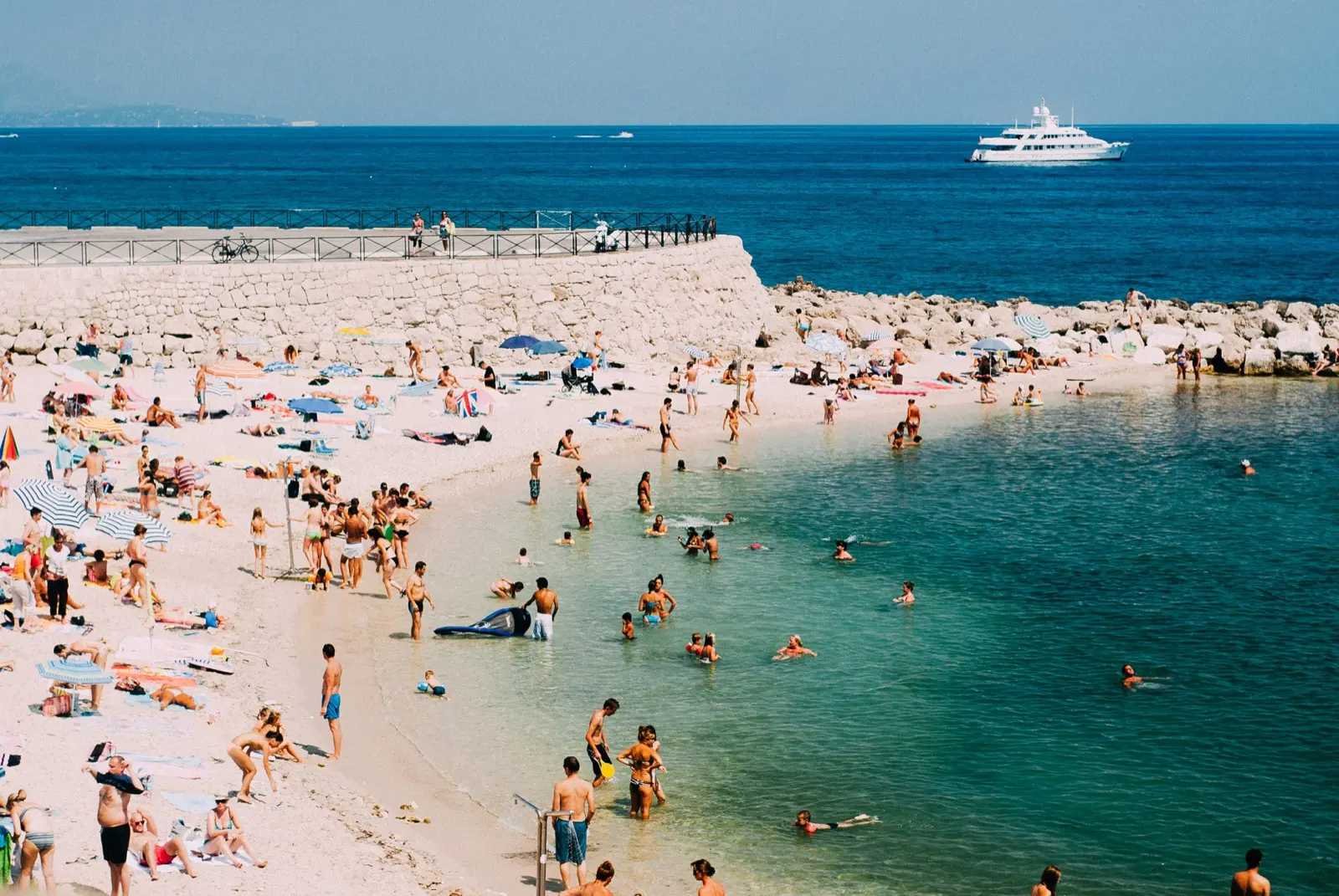 Une plage de galets très fréquentée au bord de la Méditerranée, avec des baigneurs dans une eau turquoise, des parasols rayés, une digue en pierre et un yacht blanc au large.