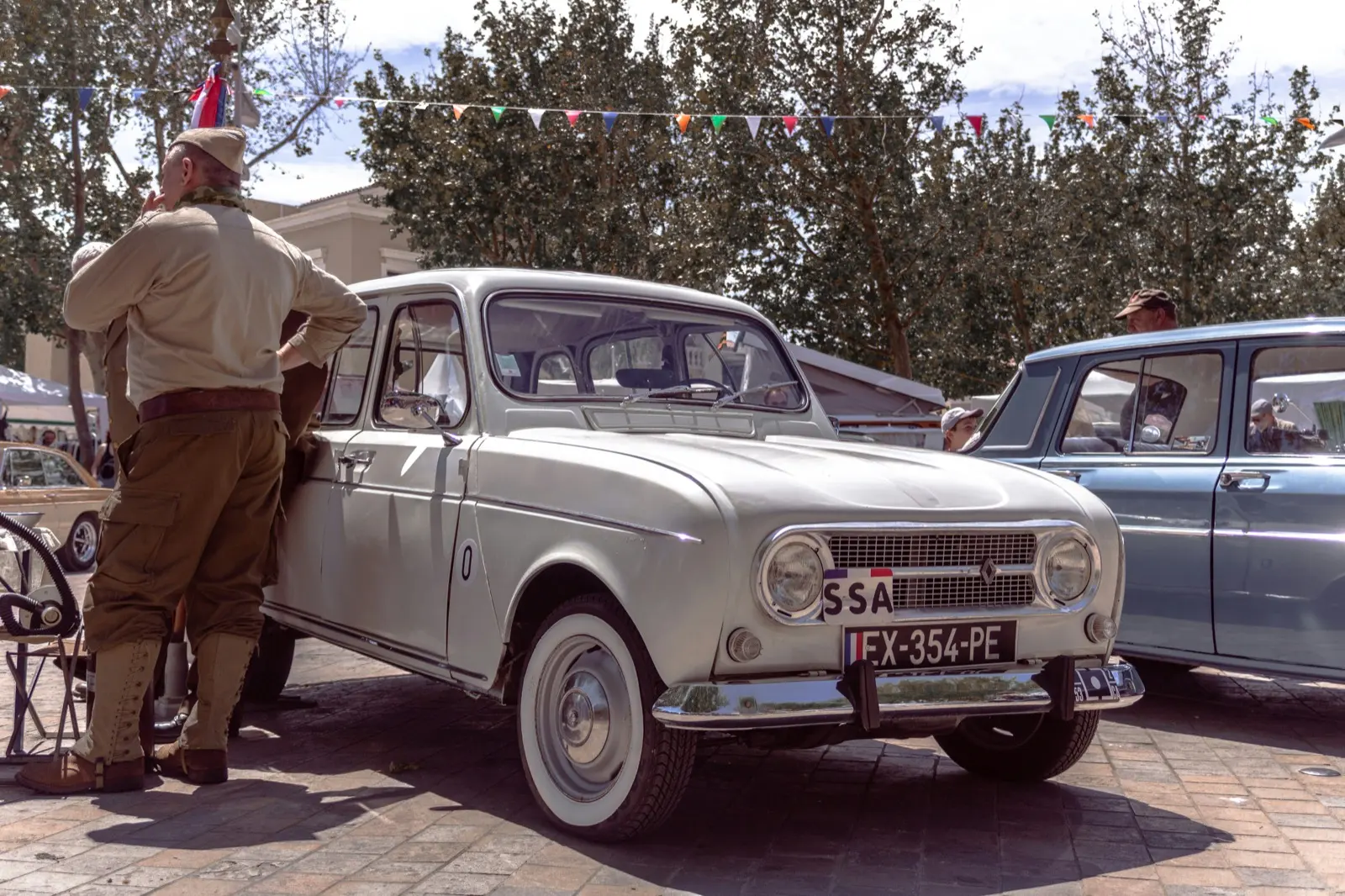 Une Renault 4 blanche de collection garée sur une place ombragée lors d’une fête de village, sous des guirlandes de fanions, à côté d’un homme en uniforme militaire d’époque.