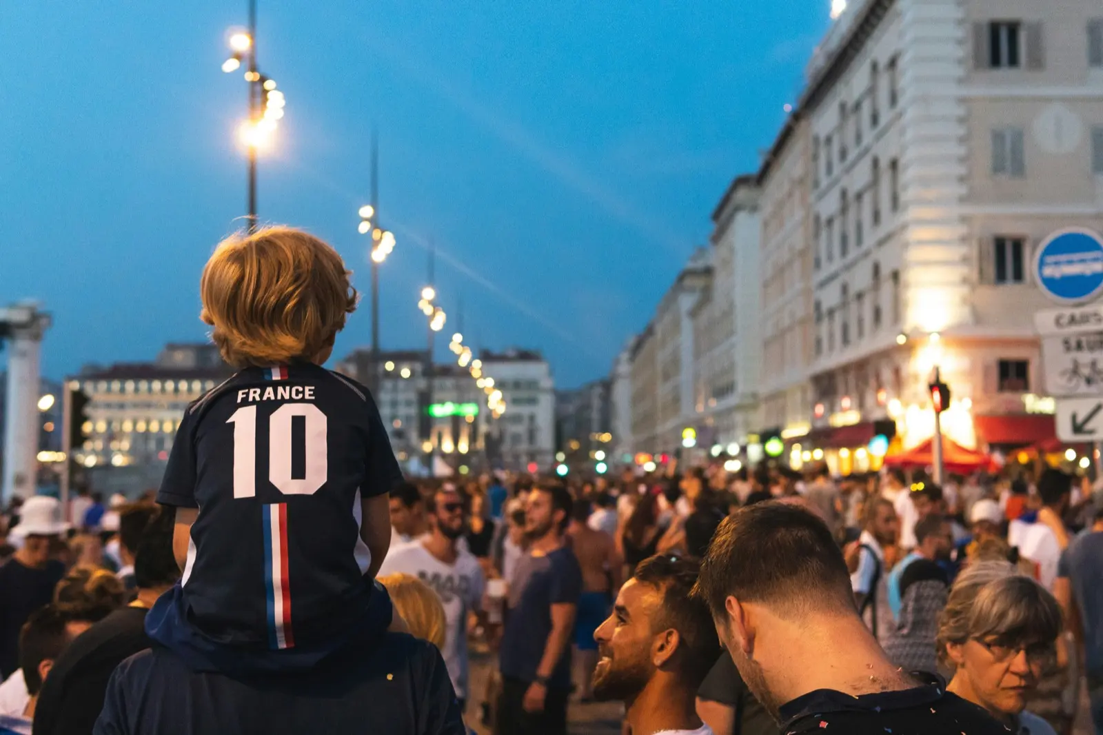 À la tombée de la nuit, un jeune enfant portant un maillot de l’équipe de France numéro 10 est assis sur les épaules d’un adulte, au milieu d’une foule de supporters sur le Vieux-Port de Marseille.